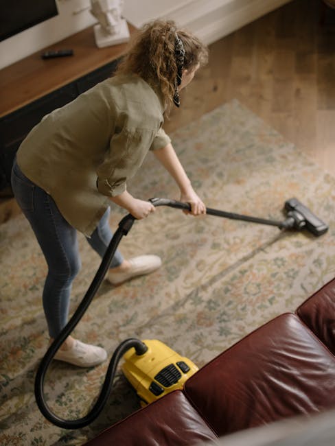 A young boy is vacuuming a beige fabric sofa in a bright living room with natural light streaming through large windows, while an older man with gray hair and a beard, dressed casually in a gray t-shi