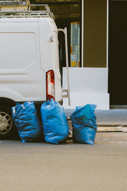 A vintage wooden sofa with a brown and orange patterned upholstery sits on a concrete pavement in front of a large pile of crumpled cardboard boxes, paper waste, and discarded packaging. The backgroun