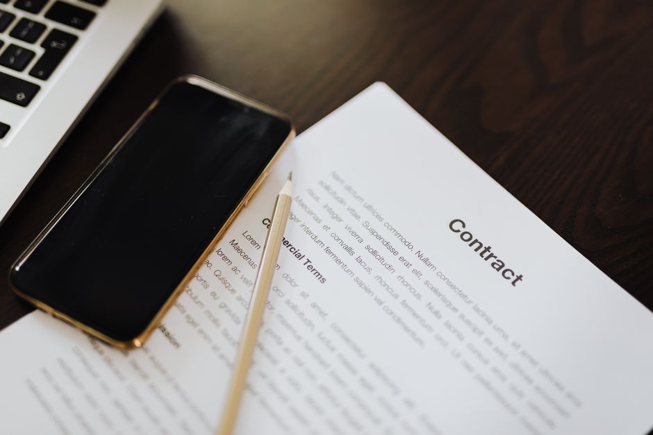Close-up view of a contract document titled 'Contract' on a dark wooden desk, accompanied by a gold-colored smartphone, a yellow pencil, and a portion of a silver laptop keyboard visible in the top le