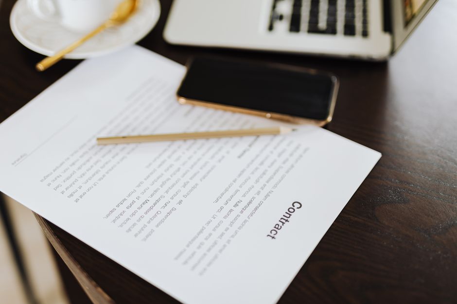 Close-up view of a contract document titled 'Contract' on a dark wooden desk, accompanied by a gold-colored smartphone, a yellow pencil, and a portion of a silver laptop keyboard visible in the top le