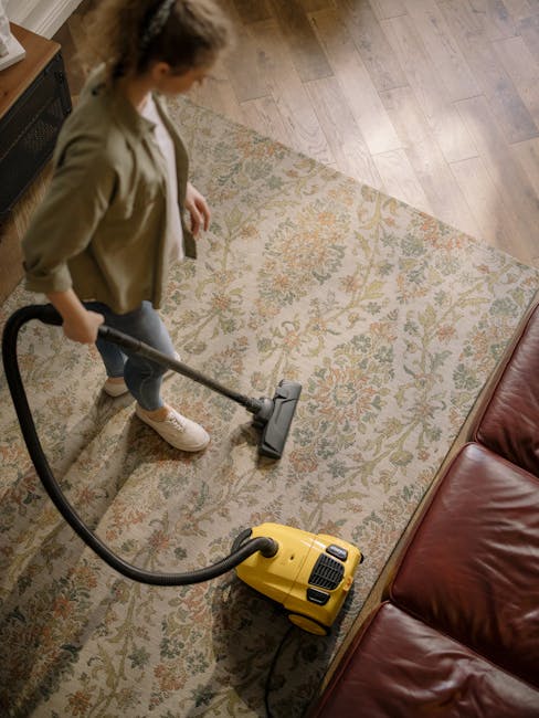 A professional cleaner from Putney Carpet Cleaners, dressed in a white protective suit and gloves, is performing deep cleaning with a steam or extraction machine on a beige carpet in a well-lit living