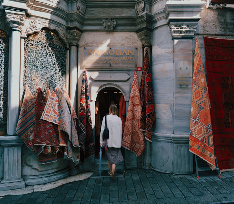 A woman enters the Anatolian shop on Putney High Street through an arched doorway framed by ornate stonework and decorative metalwork on the left, with illuminated signage reading 'Anatolian'. The exterior features patterned, colorful rugs hanging on either side of the entrance, showcasing intricate designs and warm tones. The building's facade is constructed from light-colored stone with detailed carvings, and sunlight casts shadows on the entrance area. The scene is set during daylight hours, with the woman dressed in a white coat, carrying a black bag, and walking on a paved sidewalk made of dark stones, contributing to a clean and inviting atmosphere typical of a Victorian-style commercial street. This image exemplifies the importance of surface cleaning and maintenance for creating welcoming retail environments, a service provided by Putney Carpet Cleaners for both residential and commercial spaces.