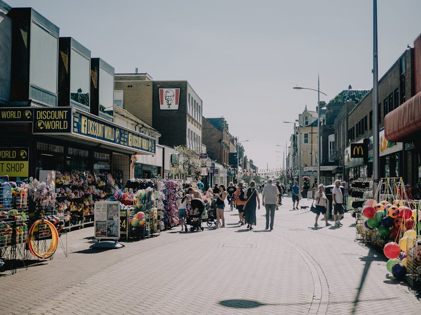 A busy outdoor shopping street on a sunny day with pedestrians walking along the pavement, lined with various retail stores including a KFC fast-food outlet and a discount shop. The storefronts feature large signage, some with bold lettering and logos. Street vendors display colorful merchandise such as balloons and toys on foldable racks and tables, creating a vibrant marketplace atmosphere. The scene is illuminated by natural daylight, casting soft shadows on the light-colored paving stones, with people of different ages casually strolling, some pushing strollers. Modern streetlights are visible along the sidewalk, and the buildings are a mix of brick and modern materials, contributing to a lively urban setting typical of a busy shopping district. This image captures the essence of a bustling retail area with diverse shopping and leisure activities, suitable for content related to surface cleaning, deep cleaning, or the general hygiene standards maintained by professional cleaning services like Putney Carpet Cleaners.