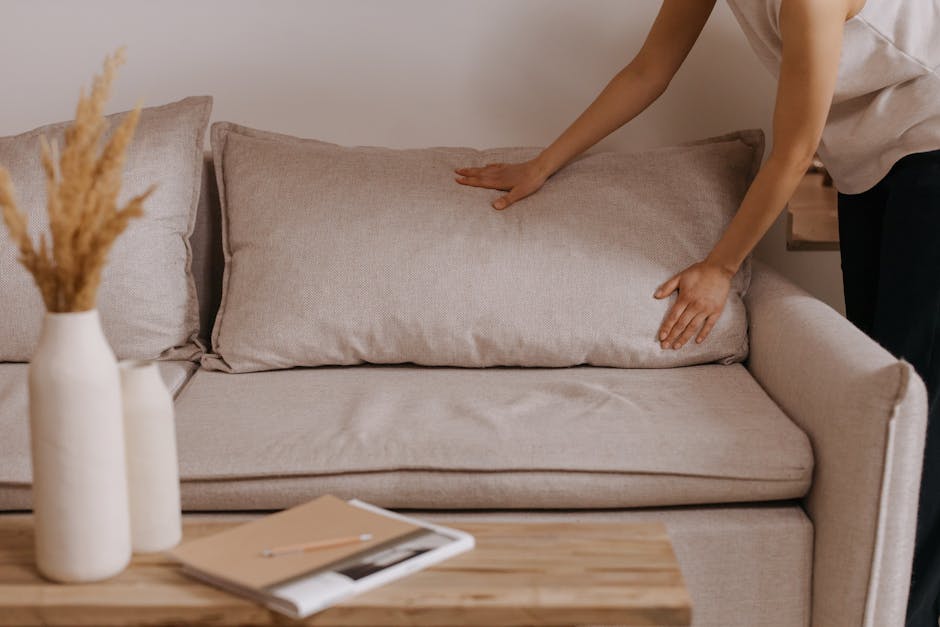 A woman with curly hair and glasses is sitting comfortably on a deep blue fabric sofa in a cozy living room, smiling while looking at her phone. The sofa is decorated with yellow and green patterned c